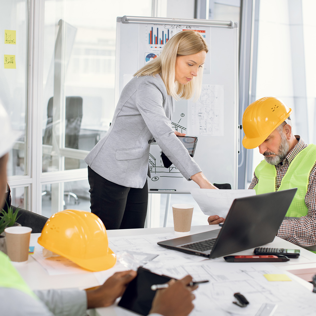 Group of coworkers in suits and helmets having business meeting at office. Responsible female engineer showing project of new building to mature male architect. Construction industry.
